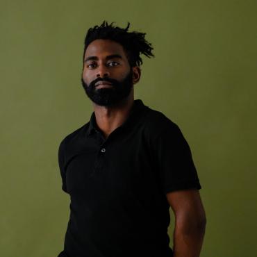 Jerron, a Black man with full beard and short locks wearing black, stands stoically in front of a green background.